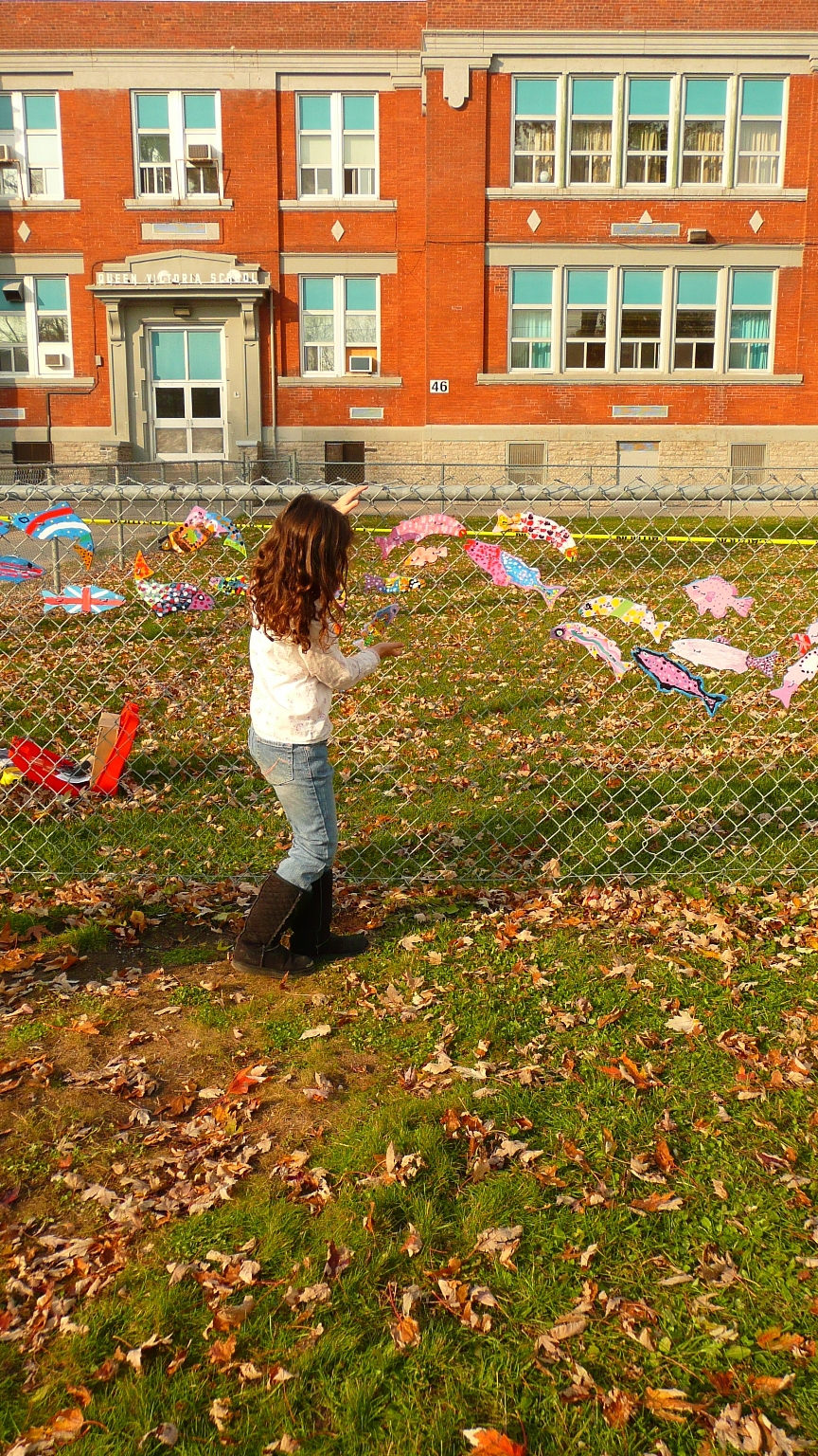 a young girl walks along a chain link fence where painted wooden fish create an art mural called a Stream of Dreams
